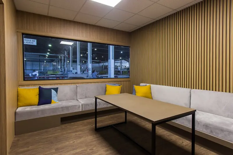 Sala de espera com sofá de canto, painel ripado e visor para a fábrica